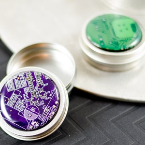 Peut inclure: Deux petits contenants métalliques ronds avec des couvercles. Les couvercles sont décorés de motifs de circuits imprimés. Un couvercle est vert et l'autre est violet.