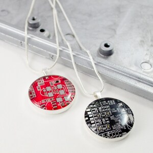 Puede incluir: Dos collares de plata con colgantes redondos que presentan patrones de placas de circuito. Un colgante es rojo con detalles plateados, el otro es negro con detalles plateados.