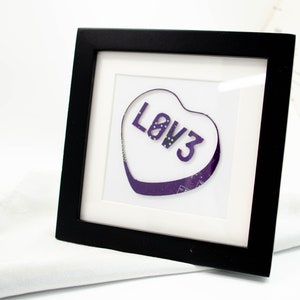 May include: A framed artwork featuring a purple heart-shaped sweet with the word "LOVE" printed on it.