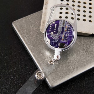 以下が含まれることがあります： 紫色の回路基板のデザインが施されたシルバーの伸縮式バッジホルダー。ホルダーには金属製のクリップと透明なプラスチック製のストラップが付いています。