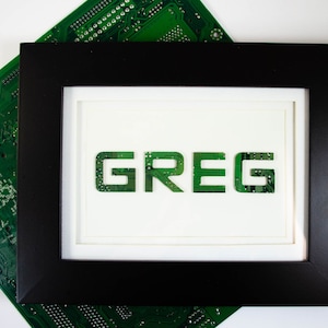 May include: A framed artwork displaying the name "GREG" in green circuit board lettering. The black frame surrounds a white mount, with a green circuit board visible in the background.