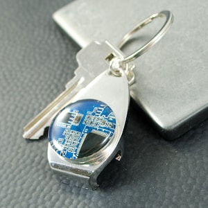 Puede incluir: Llavero plateado con un diseño de placa de circuito azul y negro en el llavero. El llavero es un abridor de botellas.