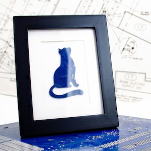 May include: A framed artwork with a blue cat silhouette made from circuit board material. The cat is set against a white background within a black frame. The artwork is displayed on a blue circuit board surface.