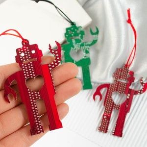 May include: Red, green, and red robot-shaped ornaments with heart cutouts. The ornaments are made from circuit board material and have small silver dots. Each robot has a string for hanging.