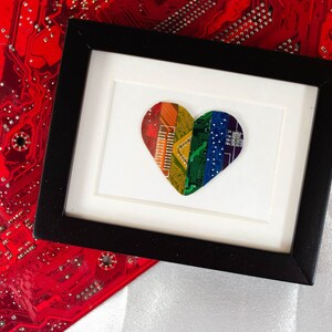 Può includere: Una cornice nera con un passepartout bianco e un cuore arcobaleno fatto di pezzi di circuito stampato all'interno. Il cuore è un simbolo di amore e tecnologia.
