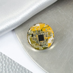 Op de afbeelding: Een ronde, gele en groene epoxyhars pin met een printplaatontwerp. De printplaat heeft een zwart vierkant in het midden.