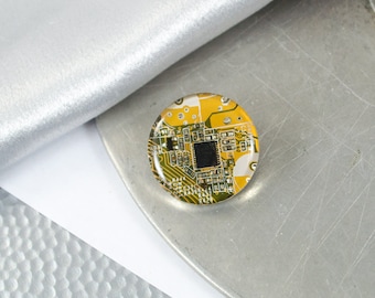 May include: A round, yellow and green, epoxy resin pin with a circuit board design. The circuit board has a black square in the centre.