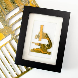 以下が含まれることがあります： 黒い額縁に白いマットが入り、その中に金色の回路基板が顕微鏡の形に作られています。回路基板は白い背景の上に置かれています。