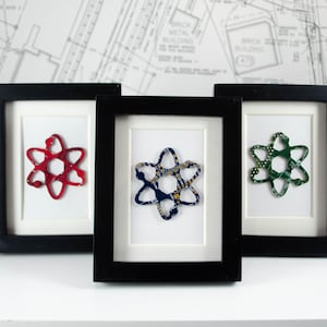 Puede incluir: Tres piezas de arte enmarcadas que presentan una forma de átomo estilizada hecha de placas de circuito recicladas. Los átomos son rojo, azul y verde.