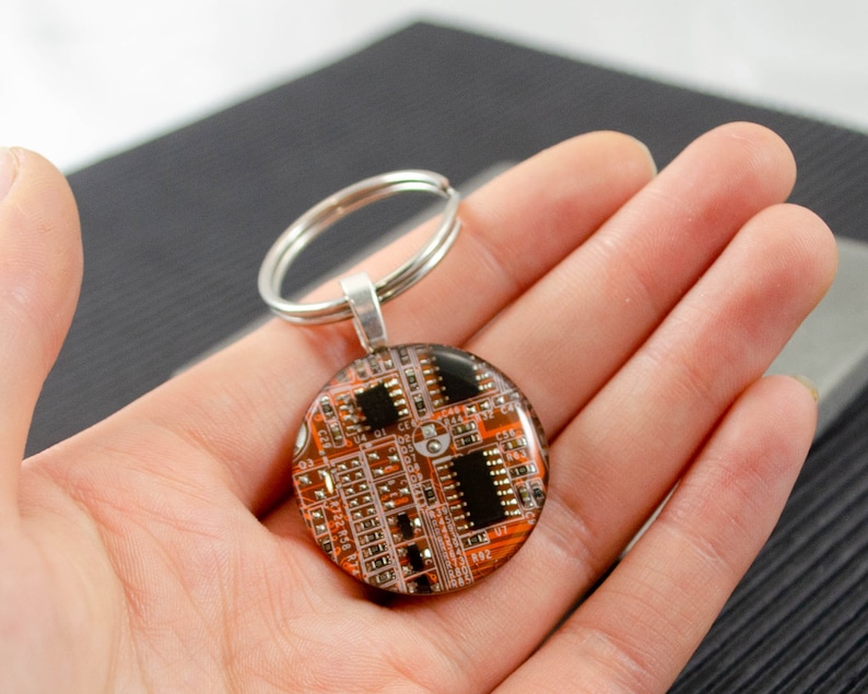May include: A round keychain with a silver key ring. The keychain features a circular resin piece with a printed image of a circuit board. The circuit board is orange and black.