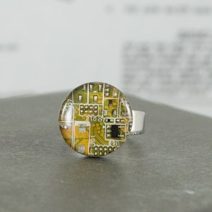 Puede incluir: Un anillo de plata con una parte superior redonda de vidrio transparente que presenta un diseño de placa de circuito verde y amarillo.