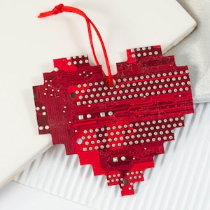 May include: A red heart-shaped ornament made from a circuit board. The ornament has a pixelated design and a red string for hanging. The circuit board is covered in silver dots and lines.