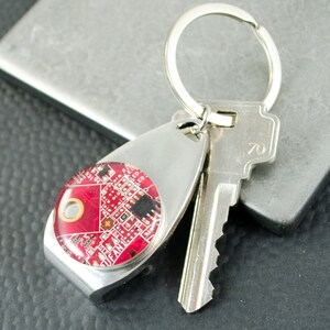 May include: A silver key ring with a red circuit board design. The key ring has a bottle opener attached to it.