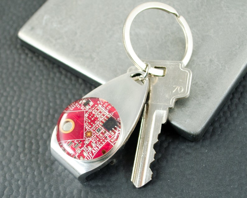 May include: A silver key ring with a red circuit board design. The key ring has a bottle opener attached to it.