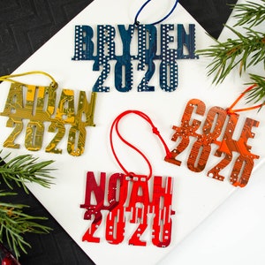 May include: Four personalized Christmas ornaments with names and the year 2020. The ornaments are in various colors, including blue, gold, red, and orange. Each ornament has a unique design and is strung with a matching colored cord.