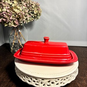 May include: A rectangular red ceramic butter dish with a matching lid featuring a small knob handle. The dish has a smooth, glossy finish and a tiered base design, sitting on a white decorative pedestal stand with a scalloped edge detail.