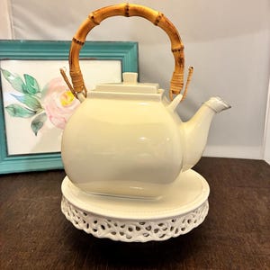 May include: Cream-colored ceramic teapot with a bamboo handle. The teapot sits on a white, decorative, round pedestal. A watercolor painting in a teal frame is in the background.
