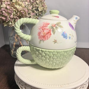 May include: A ceramic tea for one set featuring a white teapot with a floral pattern and a light green basket-weave textured cup. The teapot has a light green lid and a twisted rope-style handle, while the cup also features a matching rope handle.
