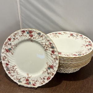 May include: A set of cream-colored porcelain plates with a floral pattern in red, blue, and brown. The plates have a gold rim and a decorative edge. One plate is on a white stand, and the others are stacked.