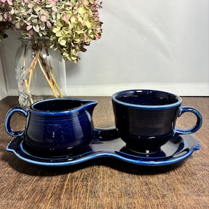 May include: A cobalt blue Fiesta cup and creamer set. The cup and creamer sit on a matching tray. The creamer has a handle and a spout. The cup has a handle. The set is on a wooden surface.
