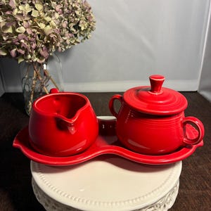 May include: A bright red ceramic creamer and a lidded sugar bowl set resting on a matching red tray. The creamer features a curved spout and handle, while the round sugar bowl has two side handles and a removable lid with a central knob.