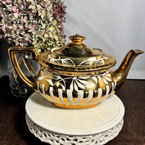 May include: A gold-colored teapot with white floral and striped designs. The teapot has a matching lid and a curved spout. It sits on a white, decorative, tiered stand. Dried purple flowers are in the background.