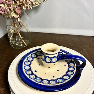May include: A ceramic candle holder with a blue and white floral design. The candle holder has a small cup for the candle and a handle. The base is a round plate with a blue border and a white center. The candle holder is on a white cake stand.