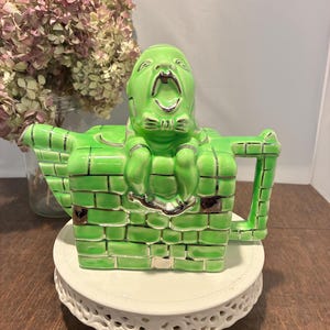 May include: A unique green ceramic teapot with a screaming face design. The teapot is shaped like a brick wall with a handle and spout. The teapot is sitting on a white, decorative cake stand. Dried flowers are in the background.