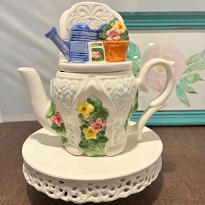 May include: A white ceramic teapot with a floral design and a matching white pedestal. The lid features a blue watering can, a small orange pot with flowers, and a small green container. The teapot has a curved handle and spout.