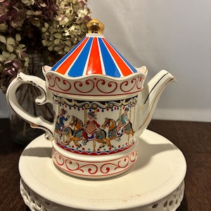 May include: A decorative ceramic teapot with a carousel design. The lid is striped red, white, and blue. The body of the teapot features carousel horses and riders. The teapot sits on a white, tiered cake stand. Gold trim accents the handle and spout.