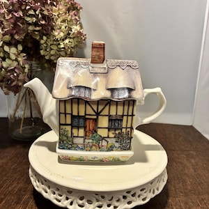 May include: A decorative teapot shaped like a cottage, featuring a thatched roof and a detailed facade with windows and a door. The teapot is cream-colored and sits on a matching, tiered, lace-edged plate. Dried flowers are in the background.