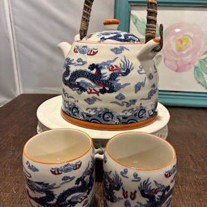 May include: A white ceramic tea set with a blue dragon design. The set includes a teapot with a woven handle and two matching cups. The rims are orange. The teapot and cups are decorated with blue dragons and clouds.