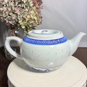 May include: White ceramic teapot with blue geometric patterns around the rim and on the lid. The teapot has a curved handle and spout. The teapot is sitting on a white decorative stand. Dried flowers are in the background.