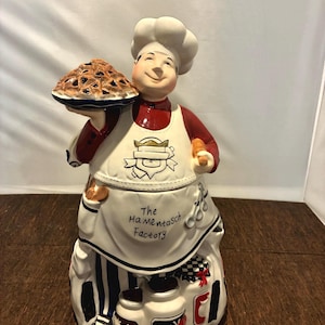 May include: A white ceramic mug with a black handle and the text "But First, Coffee" in a black, handwritten-style font. The mug is on a white surface.