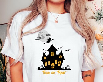 May include: White t-shirt with a black and white graphic of a haunted house, a witch flying on a broomstick, and a moon. The text "Trick or Treat" is written in yellow below the graphic.