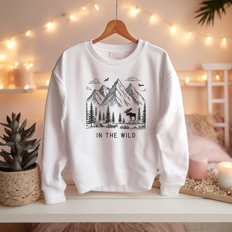 May include: White sweatshirt with a black graphic of a mountain range, a moose, and trees. The text "IN THE WILD" is printed below the graphic.