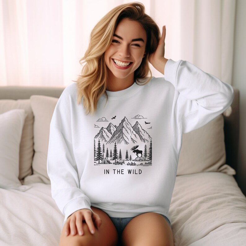 May include: White sweatshirt with a black and white graphic of a mountain range, trees, and a moose. The text "IN THE WILD" is printed below the graphic.