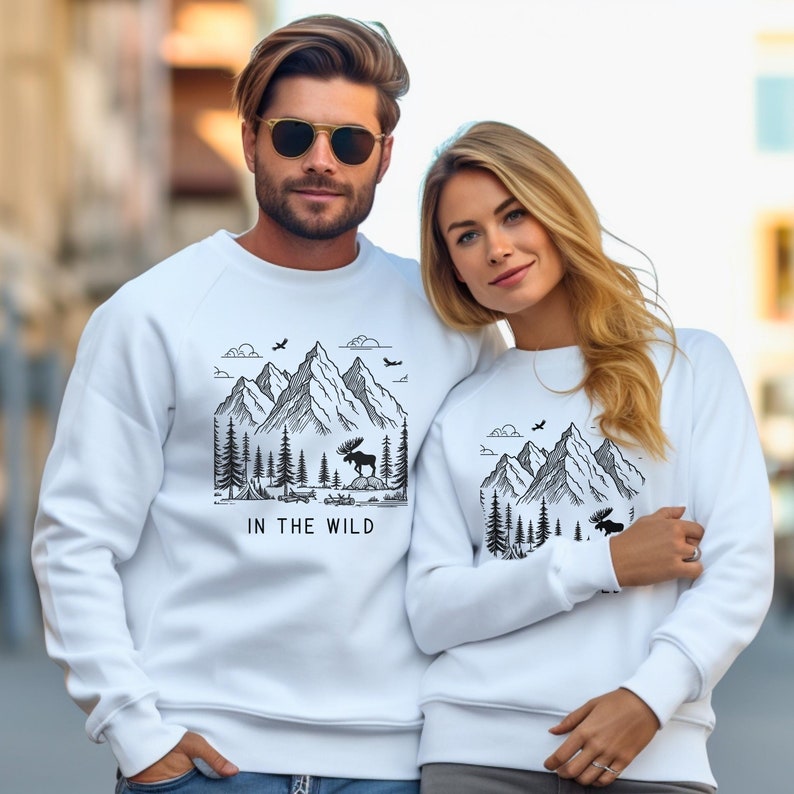 May include: White crewneck sweatshirts with a black and white graphic print of mountains, trees, and a moose. The text "IN THE WILD" is printed below the graphic.