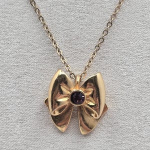 May include: Gold-toned bow pendant necklace with a small, round, purple gemstone at the center. The bow has a detailed, layered design. The chain is delicate and gold-colored. The necklace is displayed on a neutral background.