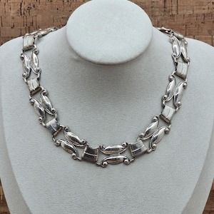 Puede incluir: Un collar plateado con un diseño geométrico repetitivo. El collar se exhibe en un busto de maniquí gris con una base de madera. Los eslabones del collar alternan entre formas rectangulares y ovaladas, creando un aspecto vintage audaz.