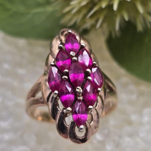 Puede incluir: Un anillo dorado con un grupo de piedras preciosas rosas vibrantes, talladas en marquesa. Las piedras están dispuestas en un patrón en cascada sobre una banda calada y detallada. El diseño del anillo es elegante y llamativo.