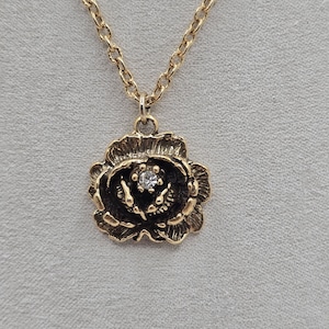 Op de afbeelding: Een goudkleurige ketting met een rozenhanger. De hanger heeft een donker centrum met een kleine, heldere edelsteen. De ketting is delicaat en ook goudkleurig, wat een klassieke en elegante uitstraling creëert. Perfect als cadeau.