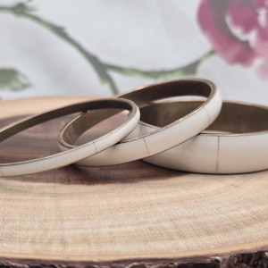 May include: Three ivory-colored bangles with brass-toned metal accents. The bangles are stacked on a wooden surface, showcasing their varying widths and segmented design. The background features a floral pattern.