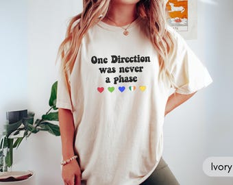 May include: Ivory-colored t-shirt with the text "One Direction was never a phase" in black. Below the text are five colorful hearts. The shirt is a casual, oversized fit.