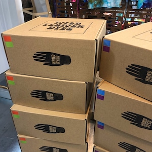 Peut inclure: Pile de six boîtes en carton brun avec le texte "WILD FLOWER DYES LIVE IN COLOR" sur la boîte du dessus. Chaque boîte a une empreinte de main noire avec le texte "HAND DYED" dessus.