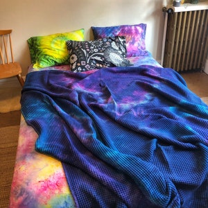 May include: A bed with tie-dye bedding in shades of blue, purple, pink, yellow, and green. The bed has a blue waffle-weave blanket and three pillows, one with a floral pattern. A wooden chair is visible in the background.