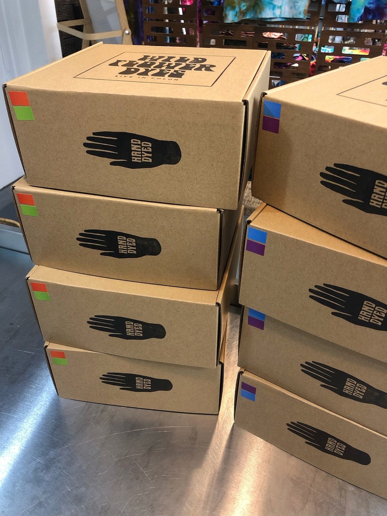 May include: Stack of six brown cardboard boxes with the text "WILD FLOWER DYES LIVE IN COLOR" on the top box. Each box has a black handprint with the text "HAND DYED" on it.