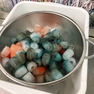 May include: A white plastic container with a metal strainer filled with colourful ice cubes. The ice cubes are various shades of blue, green, and orange.