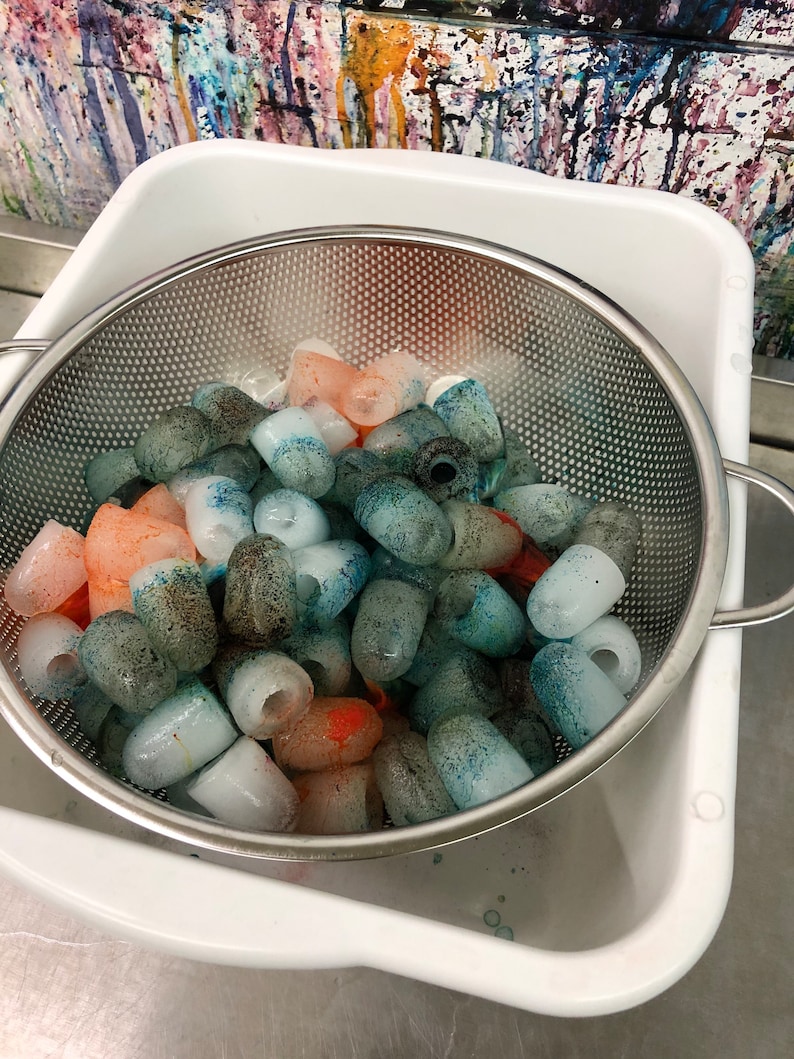 May include: A white plastic container with a metal strainer filled with colourful ice cubes. The ice cubes are various shades of blue, green, and orange.