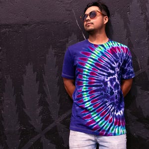 May include: A short-sleeved, tie-dye t-shirt in shades of blue, purple, green, and pink. The shirt features a spiral design and is paired with light-wash jeans. The person is wearing sunglasses and is leaning against a dark wall.